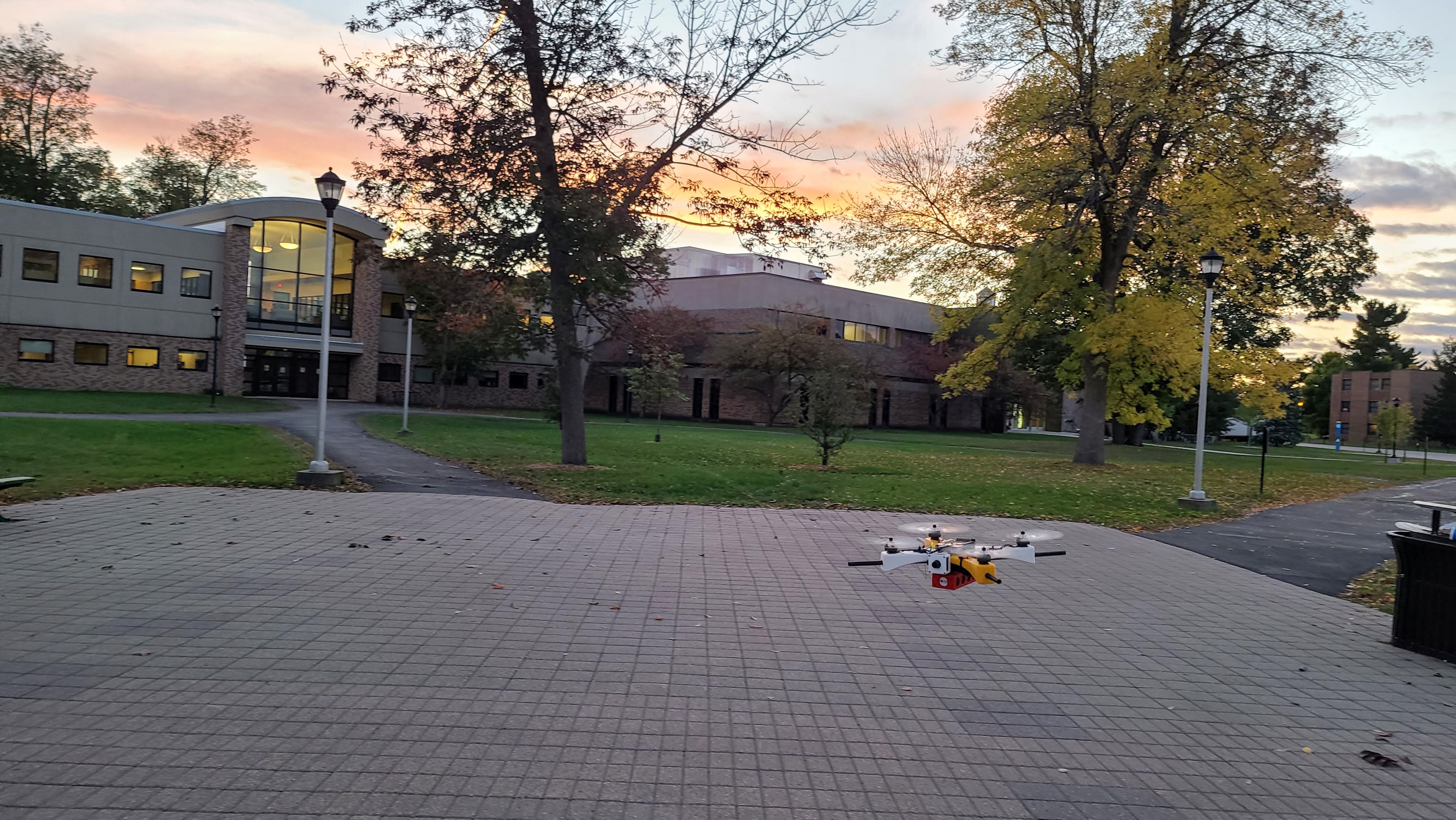 Modular quadcopter drone with yellow and red components hovering outdoors at golden hour; campus building and autumn trees in the background.