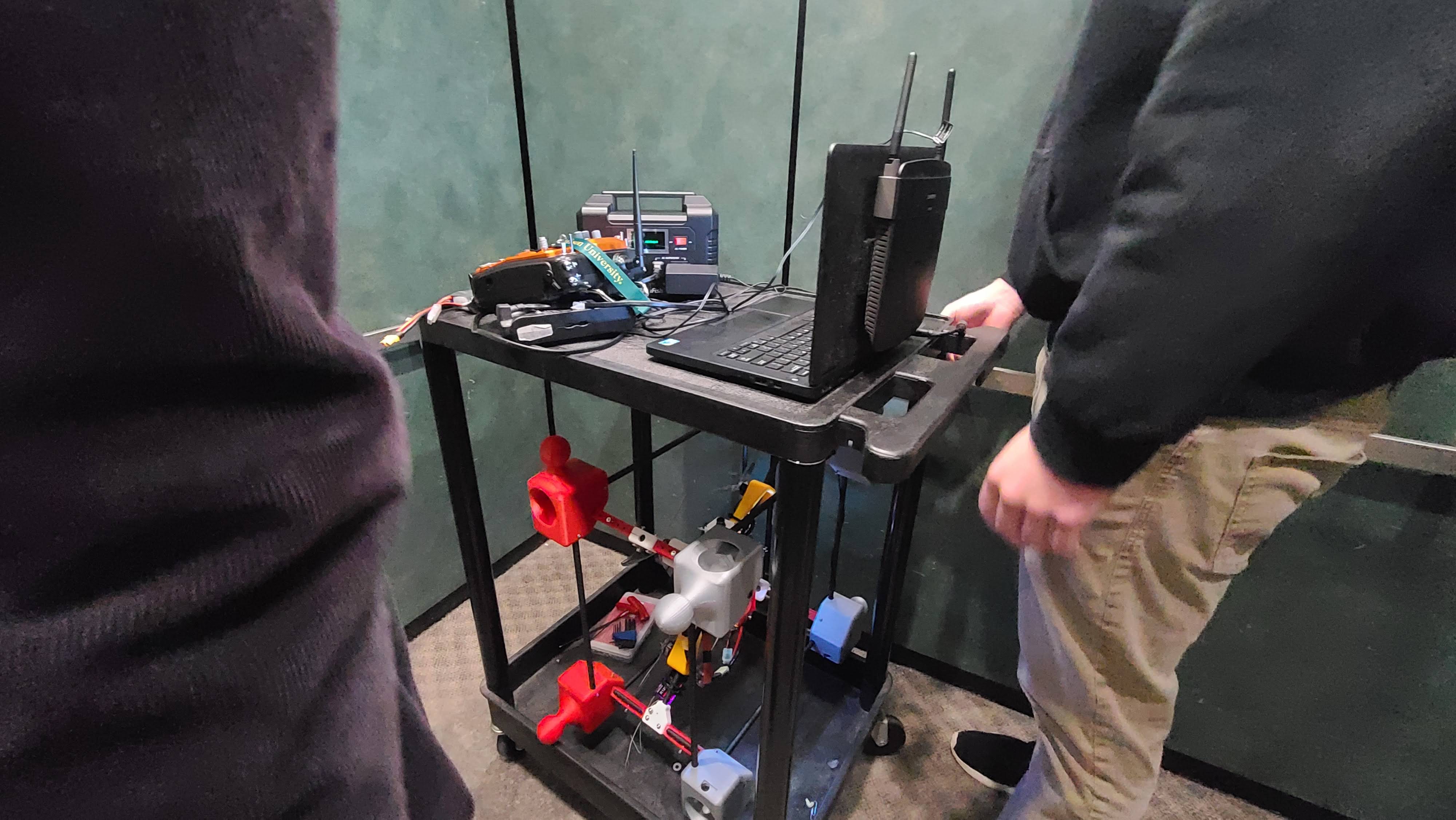 Mobile two-tier black utility cart used as a wheelable drone workstation: laptop, controller, power station, and partially assembled 3D-printed modular drone frame on the shelves.