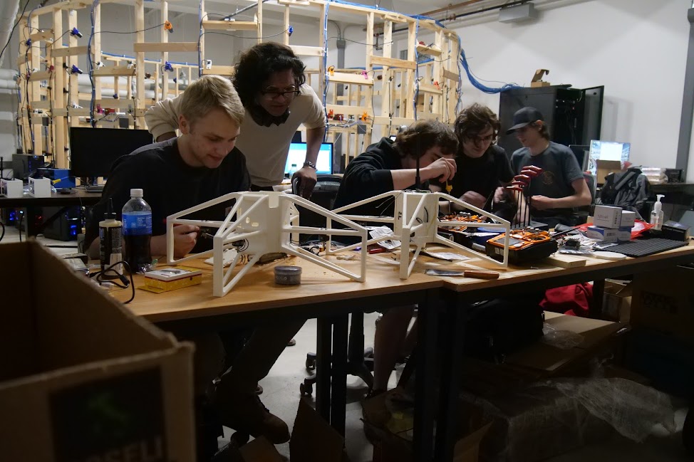 Another angle of the drone workshop: five people around a table with white modular drone frames and electronics; Alif wearing two glasses (regular and one on top of head) observing the work.
