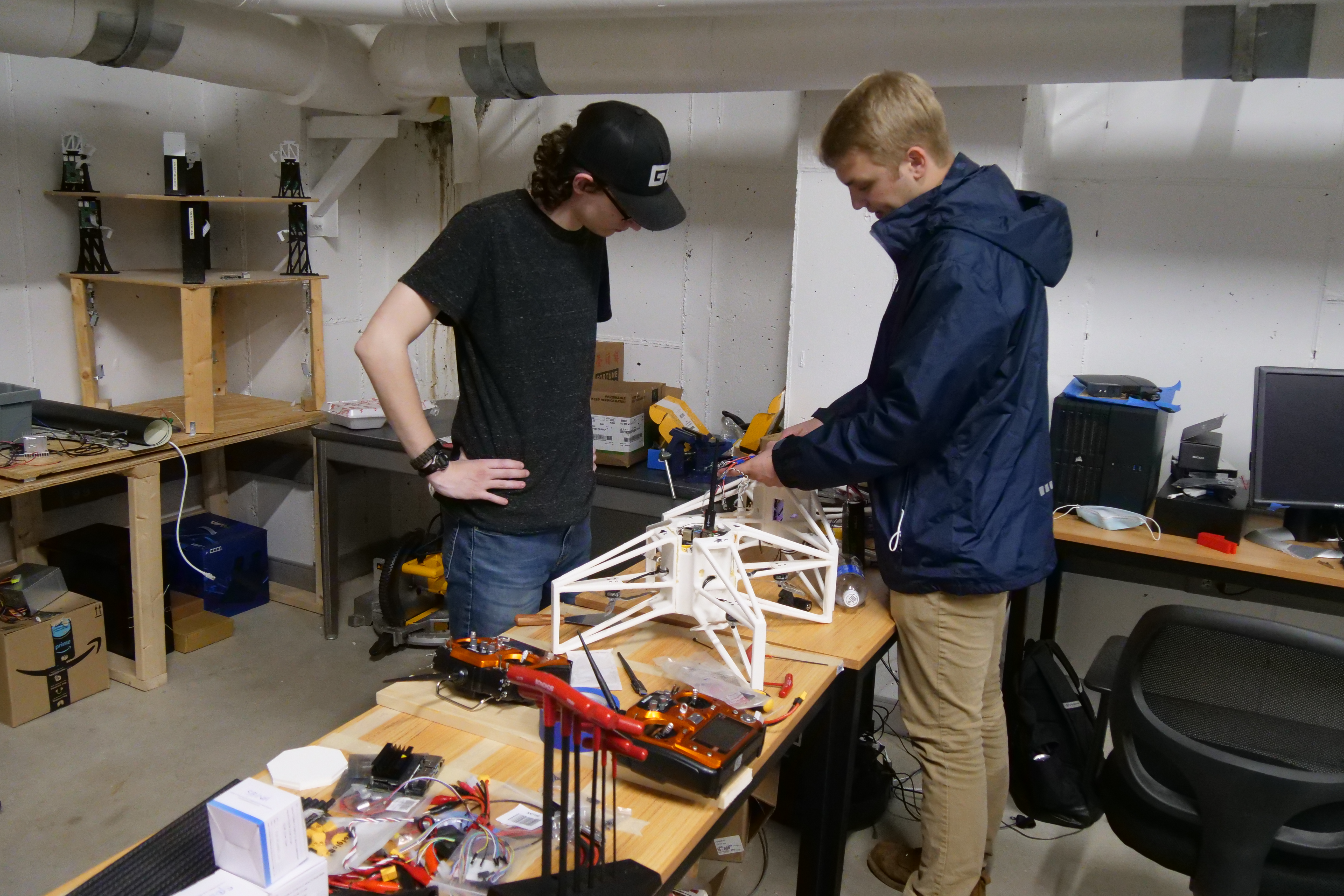 Drone workshop space: two researchers assembling a large white modular skeletal drone frame at a workbench; tools, controllers, and prototype structures in the lab.